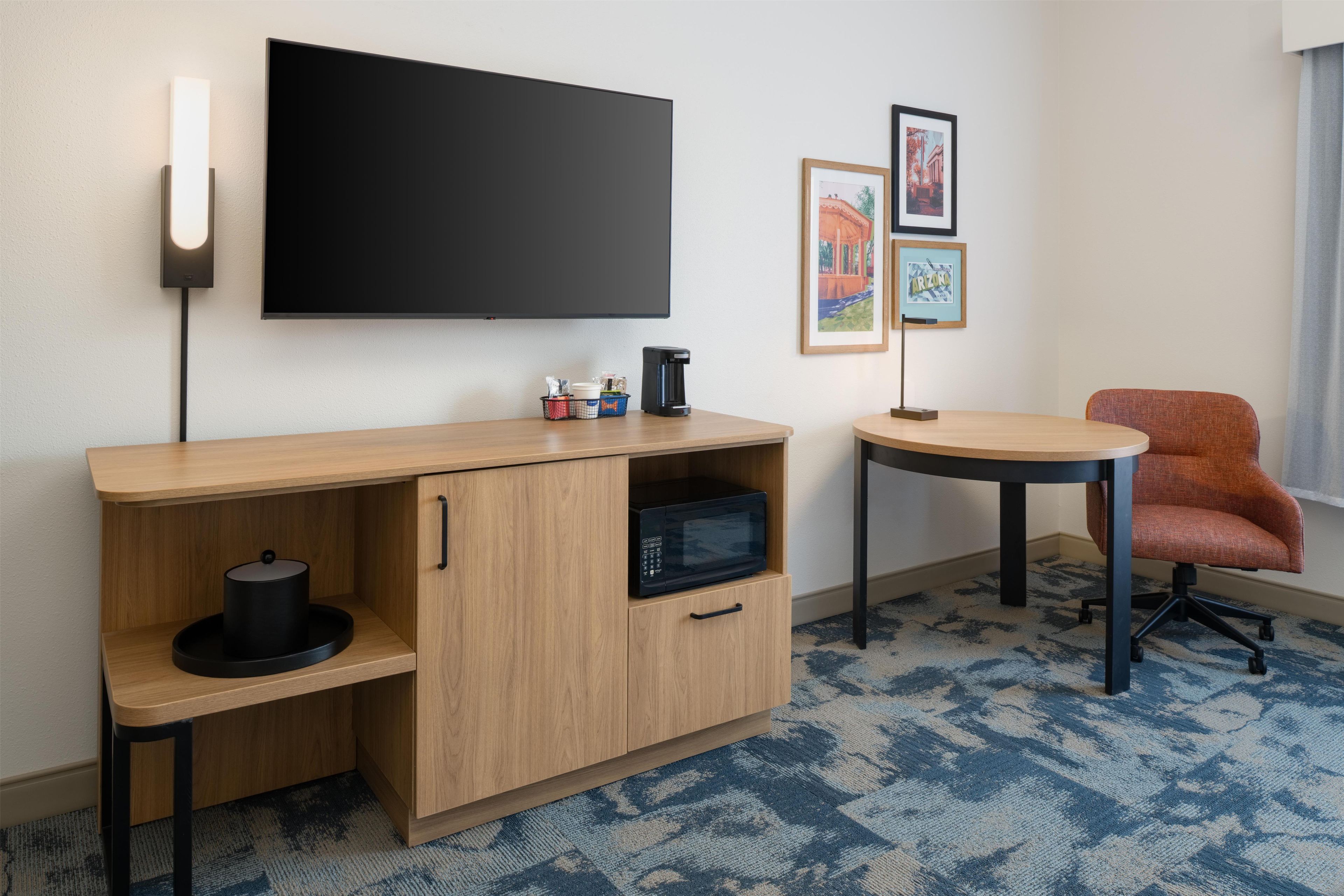 Une chambre d’hôtel équipée d’une télévision à écran plat, d’un bureau en bois avec un four à micro-ondes, une machine à café et une chaise.