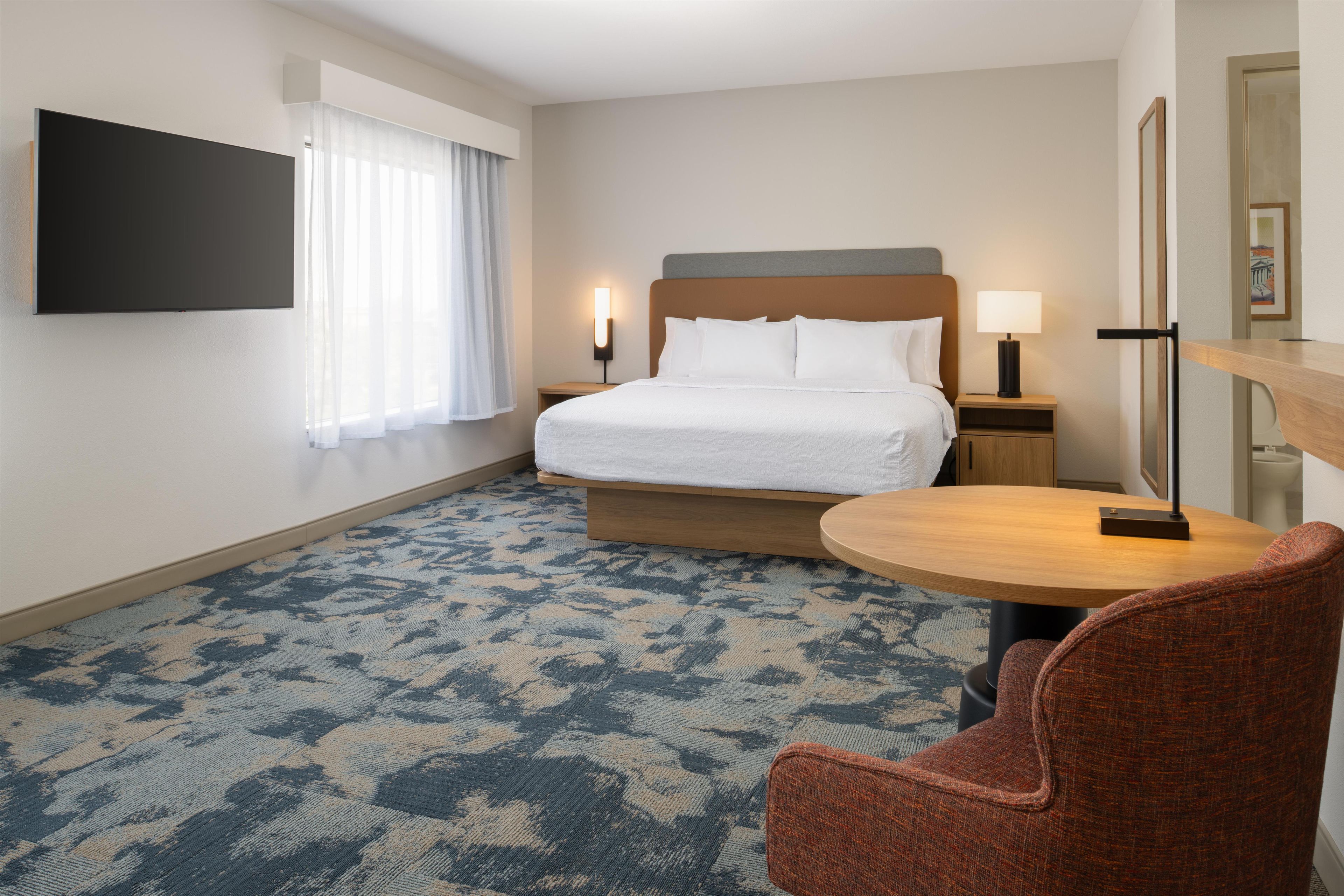 Ein Hotelzimmer mit einem Bett, einem an der Wand befestigten Fernseher, einem runden Tisch, einem Sessel und einem Nachttisch mit Lampe.