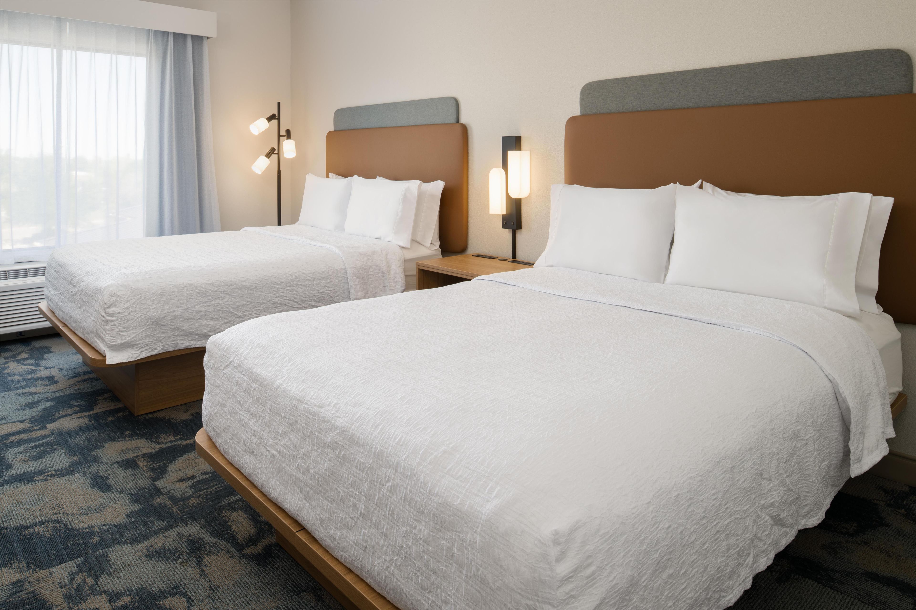 A hotel room with two beds, a nightstand with a lamp, a window with curtains, and a patterned carpet.