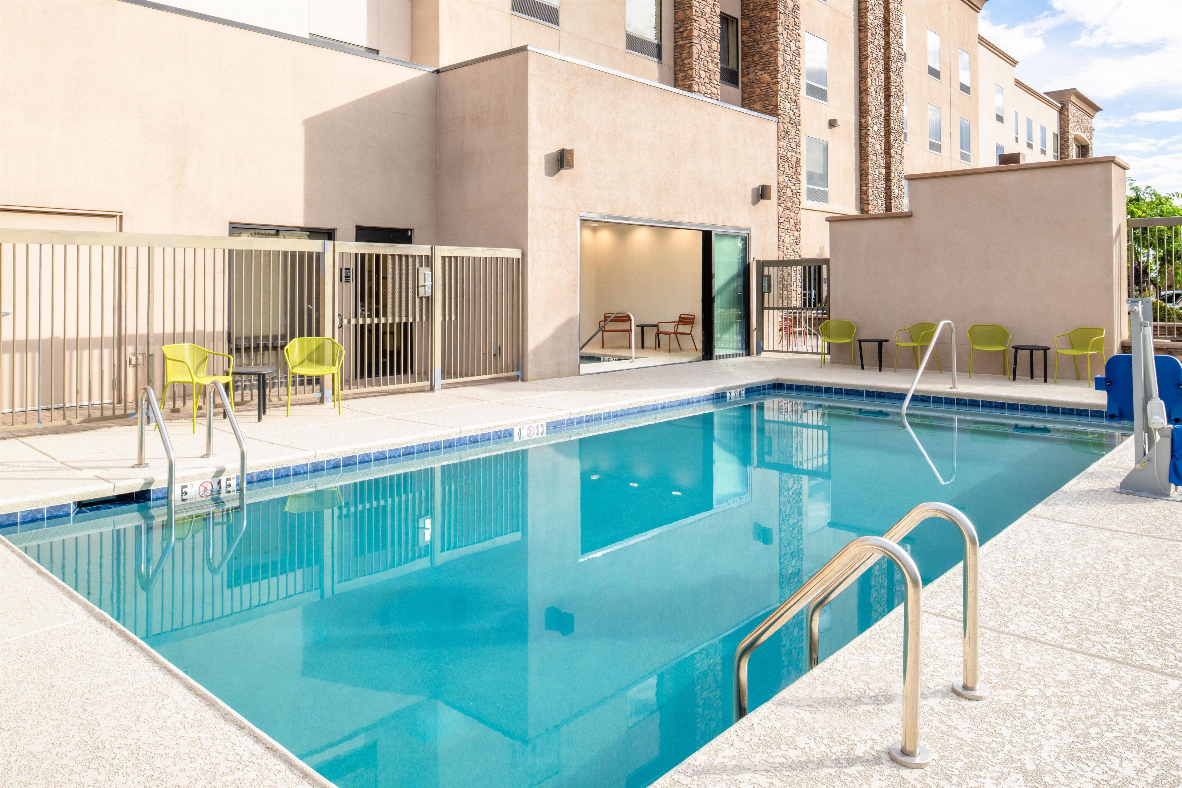 A swimming pool with a clear blue water, surrounded by a beige building with stone accents, and yellow chairs.