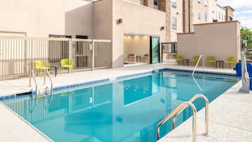 A swimming pool with a clear blue water, surrounded by a beige building with stone accents, and yellow chairs.