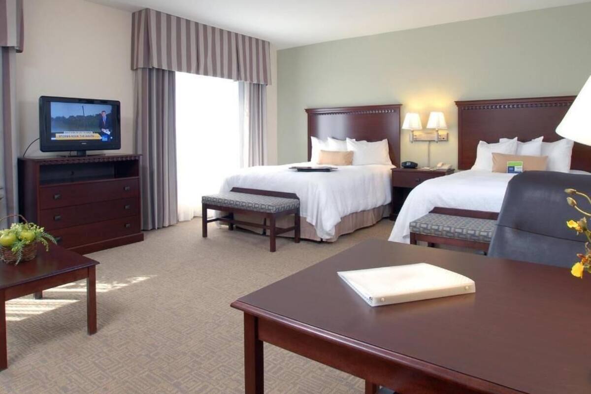 Ein Hotelzimmer mit zwei Betten, einem Schreibtisch, einem Fernseher und einem fensterbepflanzten Fenster.