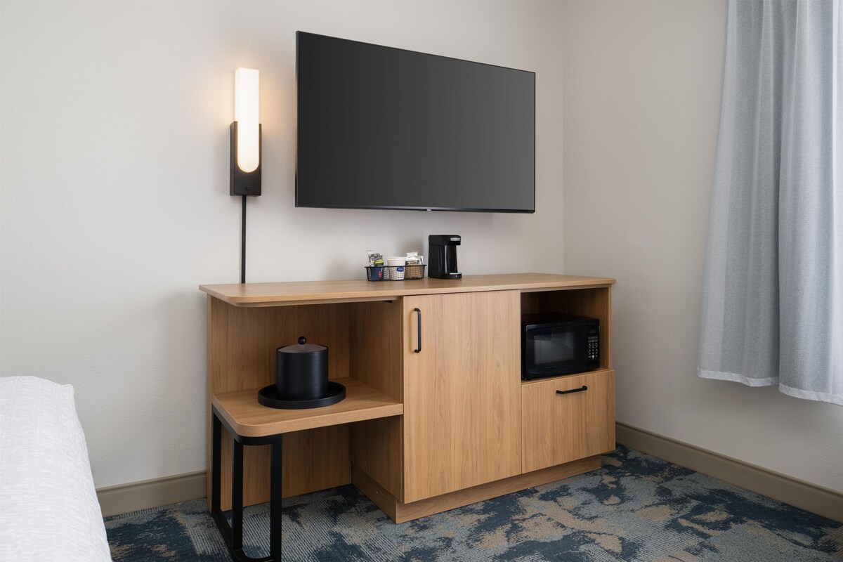 A hotel room with a flat-screen TV mounted on the wall, a wooden desk with a coffee maker, a microwave, and a small shelf with a kettle.