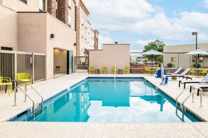 Piscina con sillas y sombrillas, rodeada de edificios.