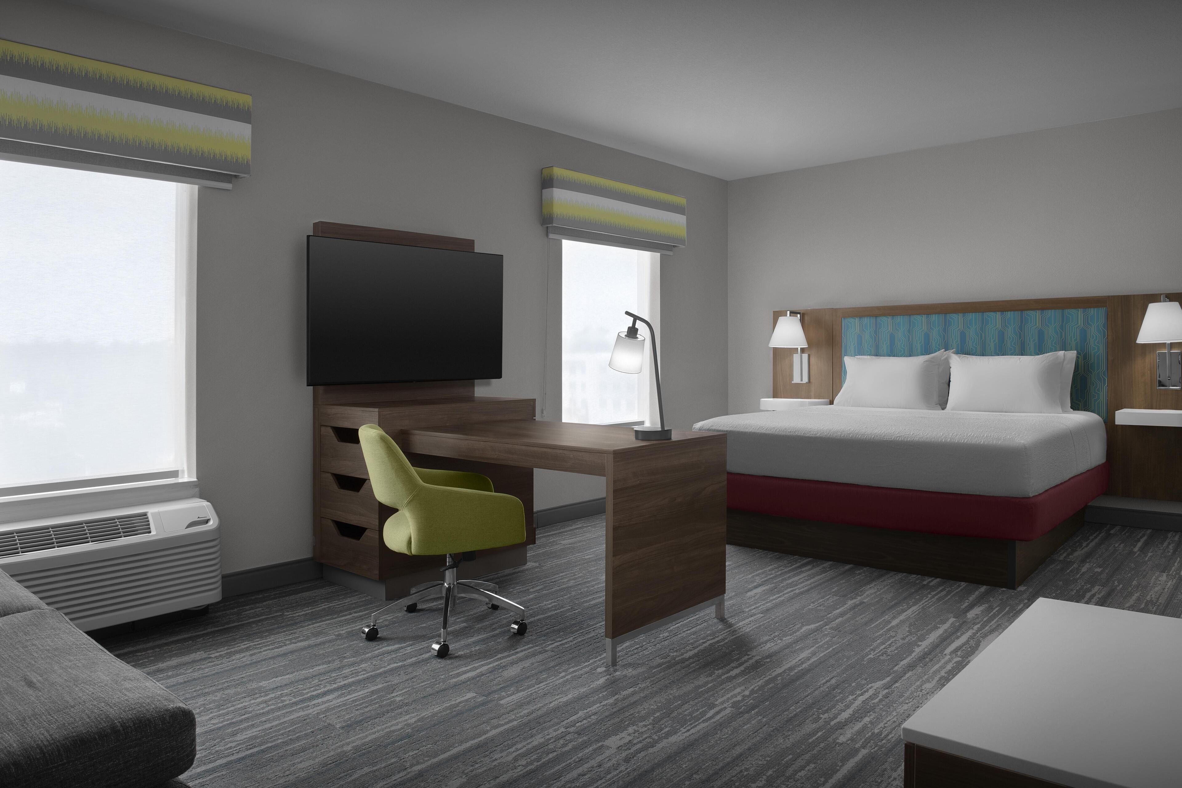 A hotel room with a bed, a desk with a chair, a television, and a window with blinds.