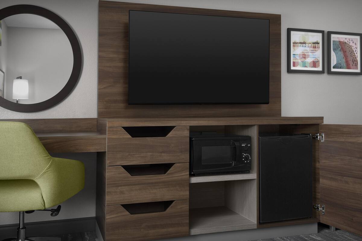 A modern hotel room with a flat-screen TV mounted above a wooden cabinet, a microwave, and a refrigerator. There is a green chair and a round mirror on the wall.