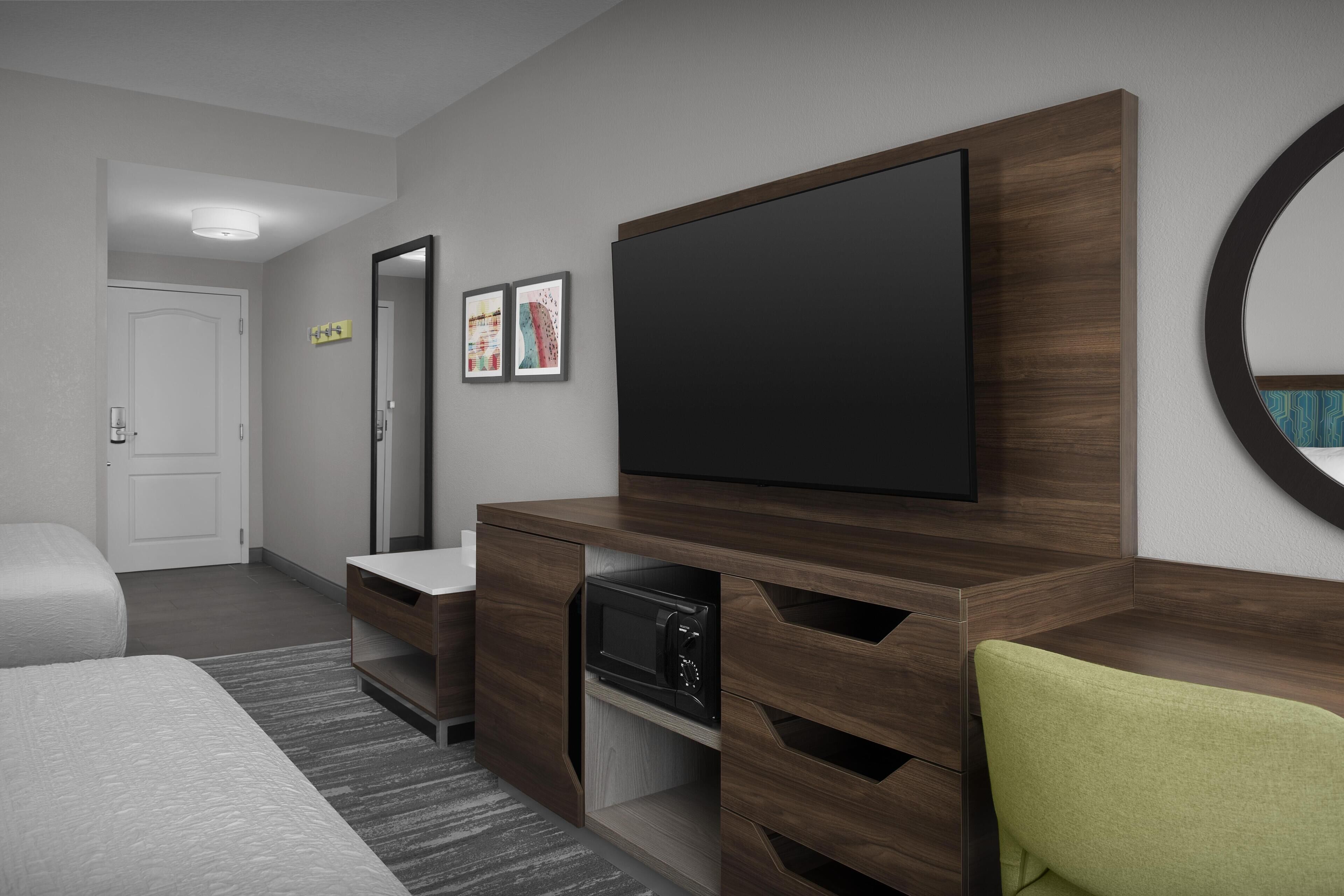 A modern hotel room with a large flat-screen TV, a microwave, a wooden cabinet, and a green chair.