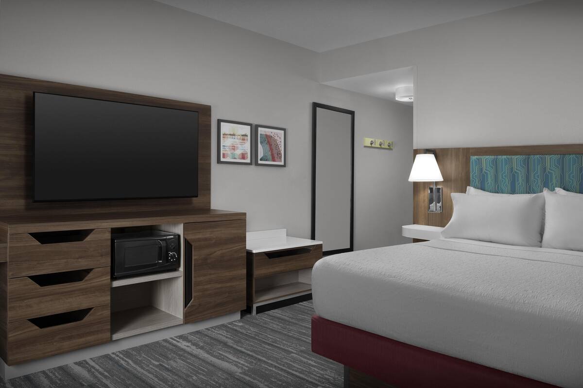 A hotel room with a large flat-screen TV, a microwave, a bedside table with a lamp, and a bed with white bedding.