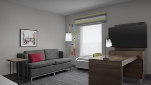 Habitación de hotel moderna con un sofá gris, una mesita, un escritorio con silla, un televisor de pantalla plana, una lámpara de pie y un ventanal con vistas.