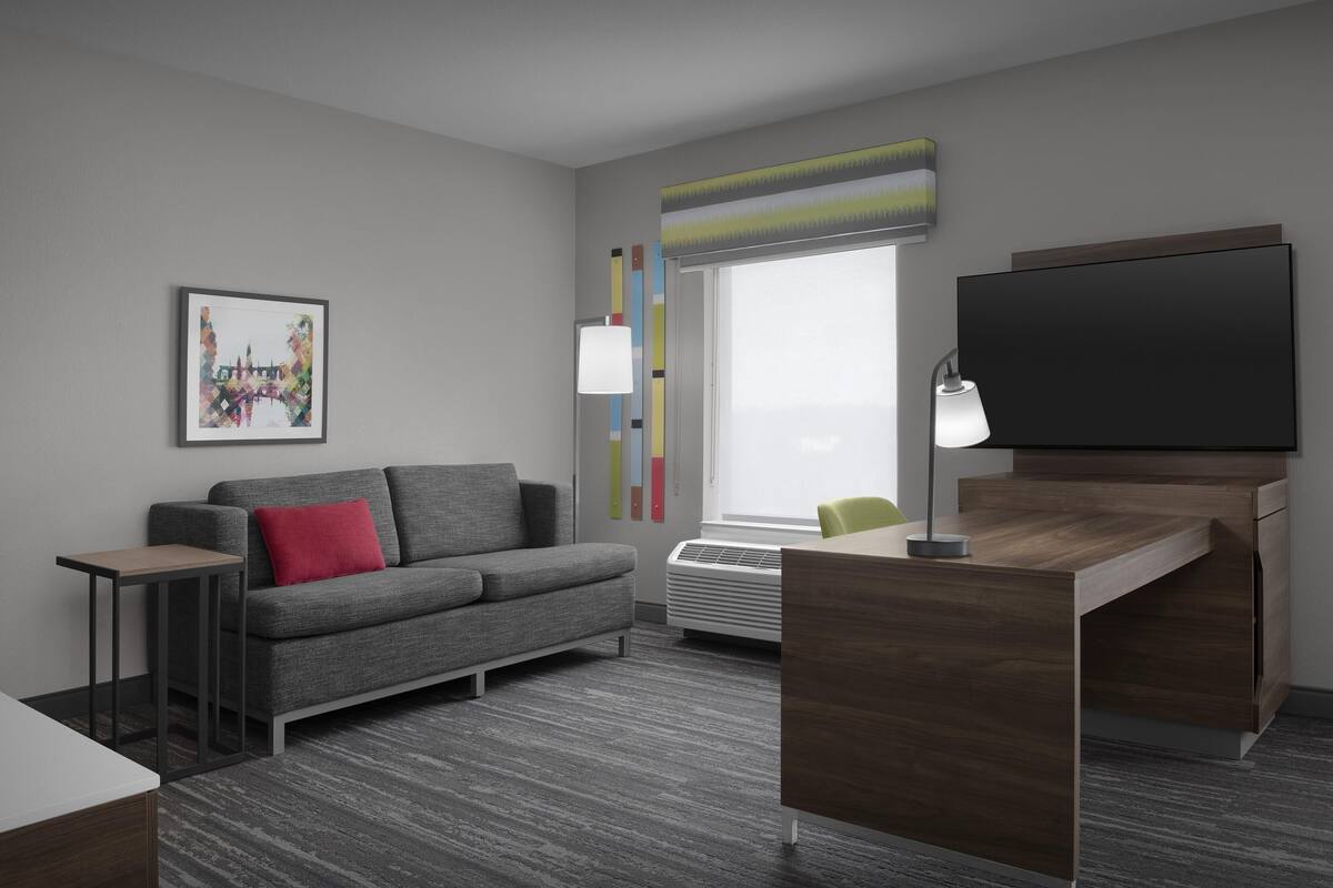 A modern hotel room with a grey sofa, a small table, a desk with a chair, a flat-screen TV, a standing lamp, and a large window with a view.