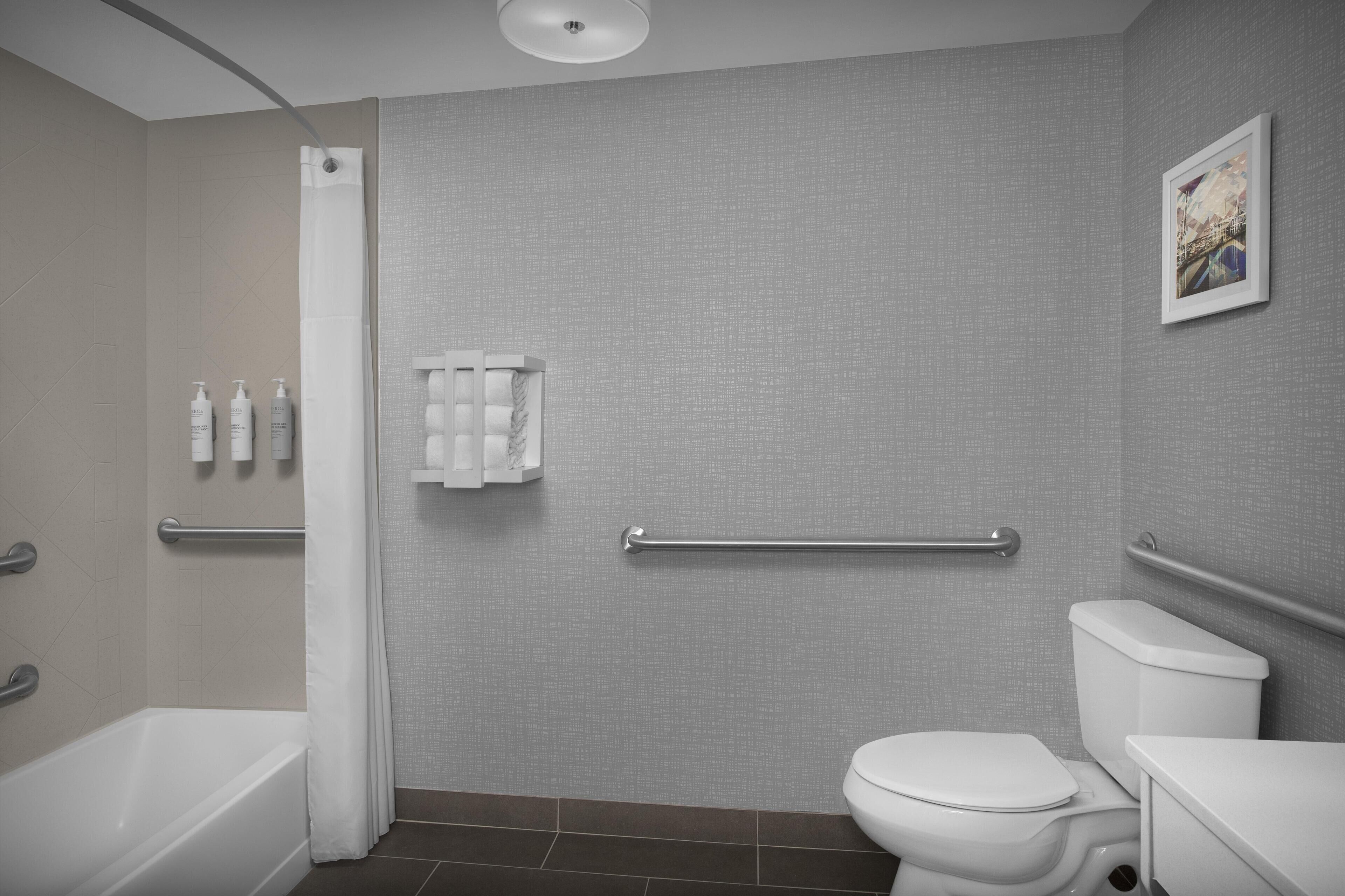 A modern bathroom with a bathtub, toilet, and shower. There are handrails and a towel rack on the wall.