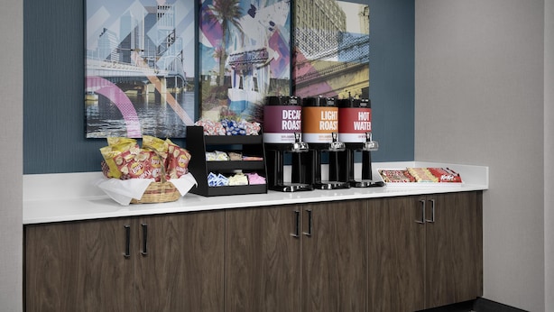 A counter with coffee dispensers, snacks, and a decorative wall with cityscape artwork. - Hampton Inn & Suites Jacksonville South - Bartram Park (Jacksonville)