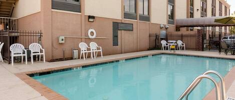 Una piscina rectangular con una escalera metálica, rodeada por una terraza de baldosas color beige. Hay sillas de plástico blanco y una mesa con un paraguas amarillo.