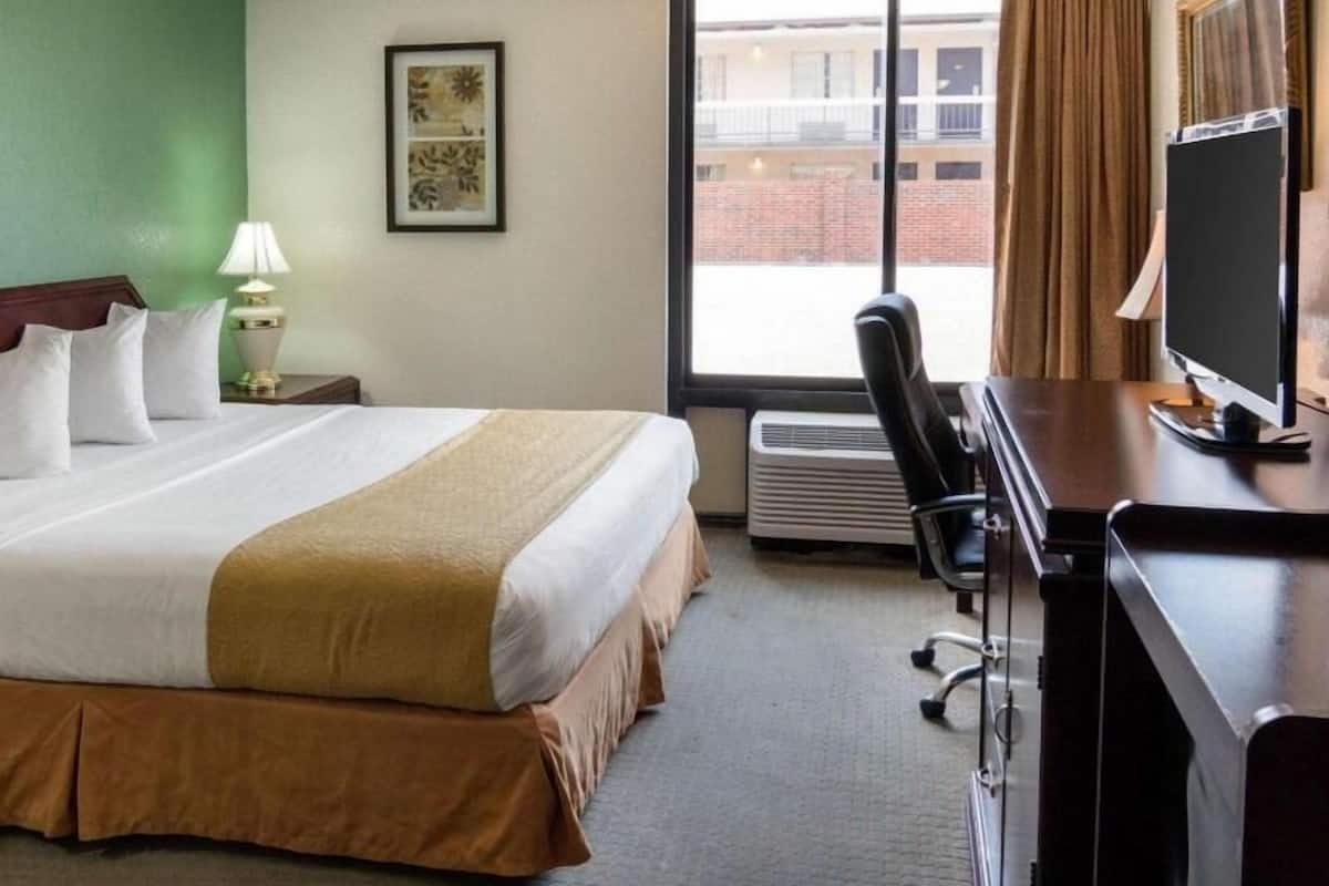 Una habitación de hotel con cama, escritorio con computadora, silla, ventana con cortinas e imagen en el pared.