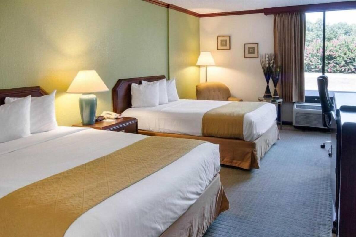 Habitación de hotel con dos camas, un escritorio, una silla, una lámpara y una ventana con cortinas.