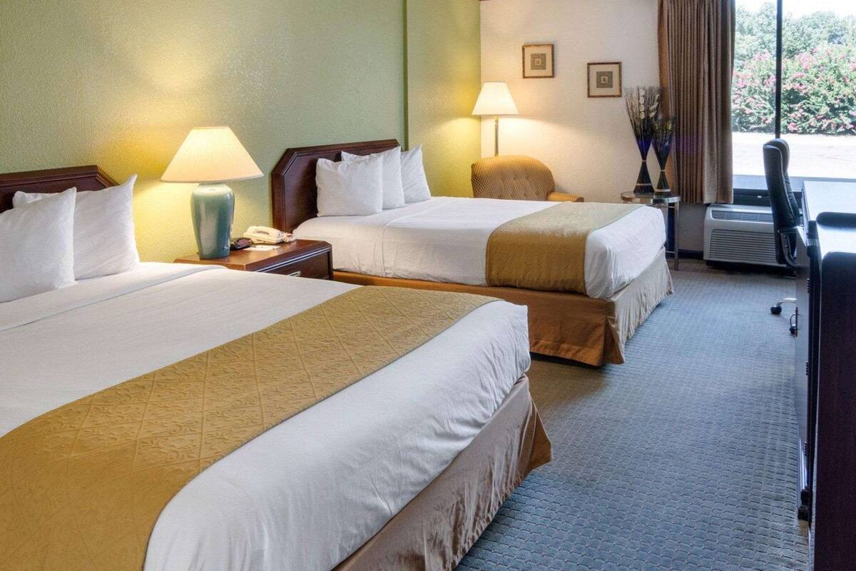 A hotel room with two beds, a desk, a chair, a lamp, and a view of the outdoors.