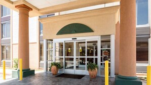 A building entrance with a glass door, two potted plants, and yellow bollards. - Quality Inn Shreveport Airport (Shreveport)