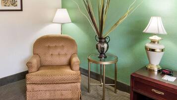 A room with a patterned armchair, a small round table with a vase of tall, thin plants, a lamp, and a wooden nightstand with a drawer.