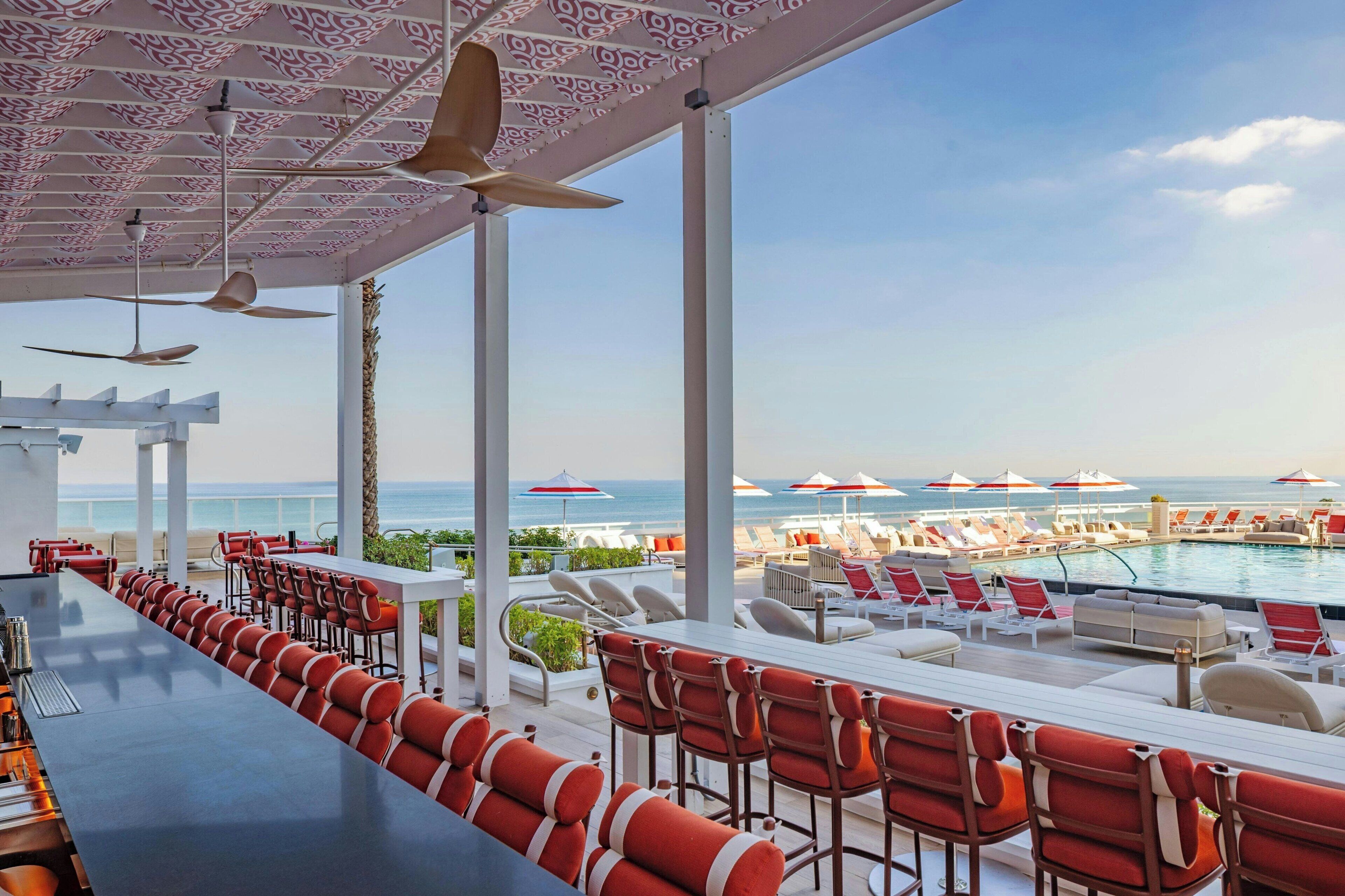 Un espace aménagé au bord de la piscine, avec un long bar, des sièges rouges et blancs et des parasols.