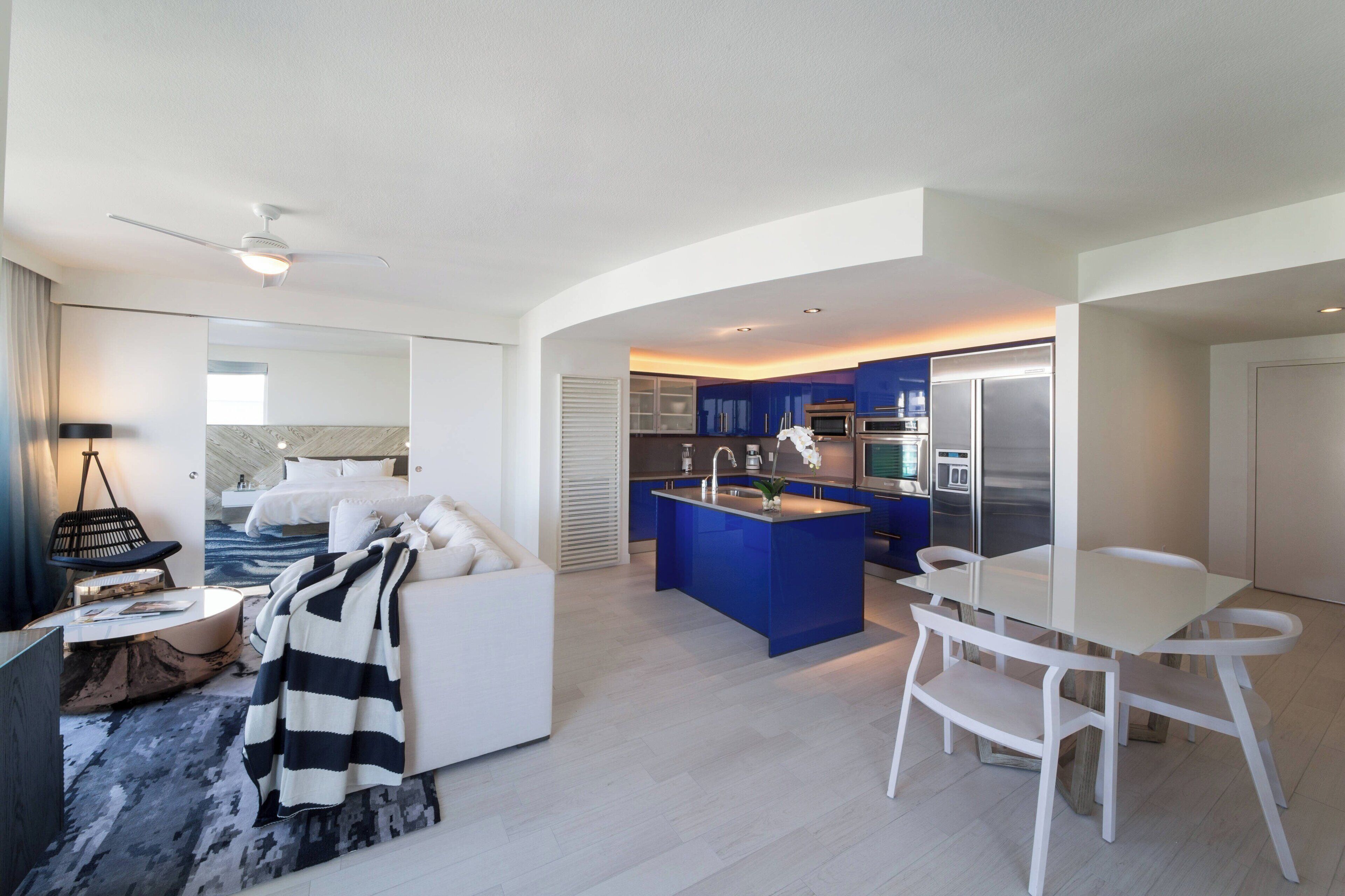 A modern hotel room with a bed, a sitting area with a sofa and a coffee table, a small kitchen area with a blue island, and a dining table with chairs.