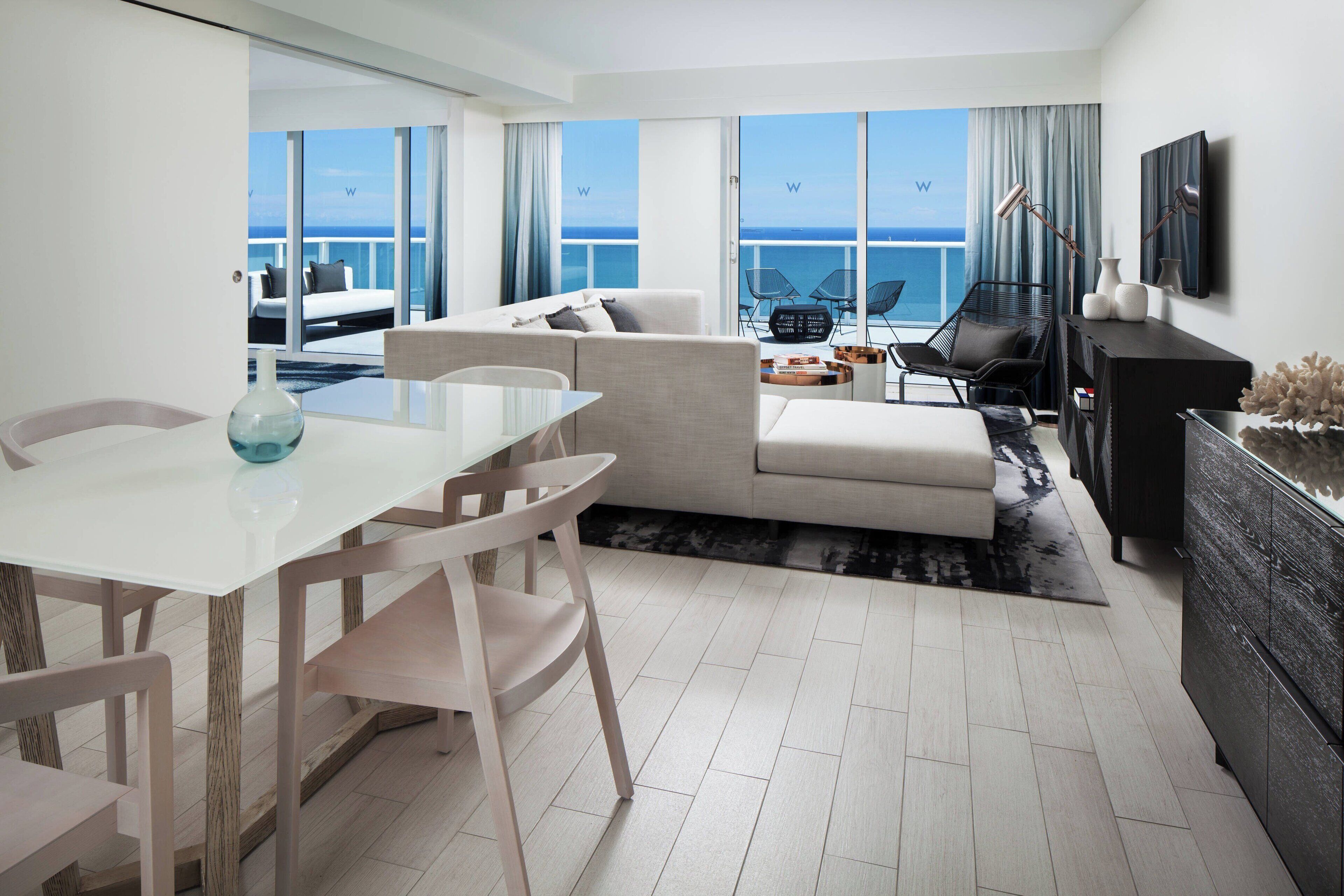 A modern living room with a white sofa, a dining table with chairs, and a view of the ocean through large windows.