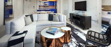 Una habitación de hotel moderna con sofá modular, una mesa de centro redonda, televisor y zona de comedor con armarios azules.