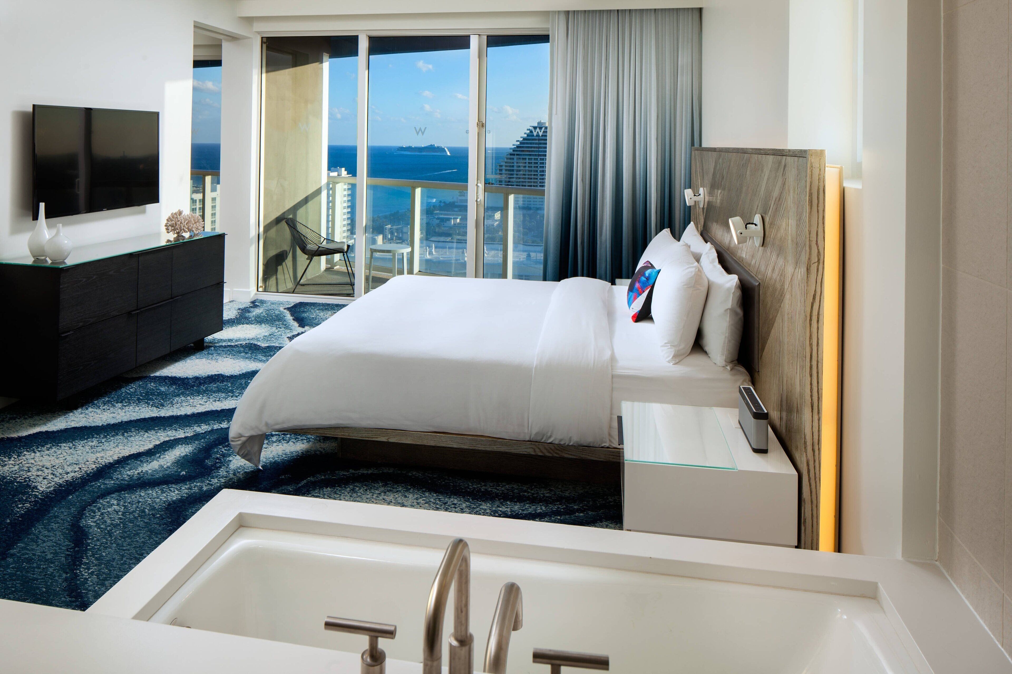 Habitación de hotel moderna con una cama grande, balcón con cerramiento de vidrio y vista a la ciudad, televisor de pantalla plana y bañera.