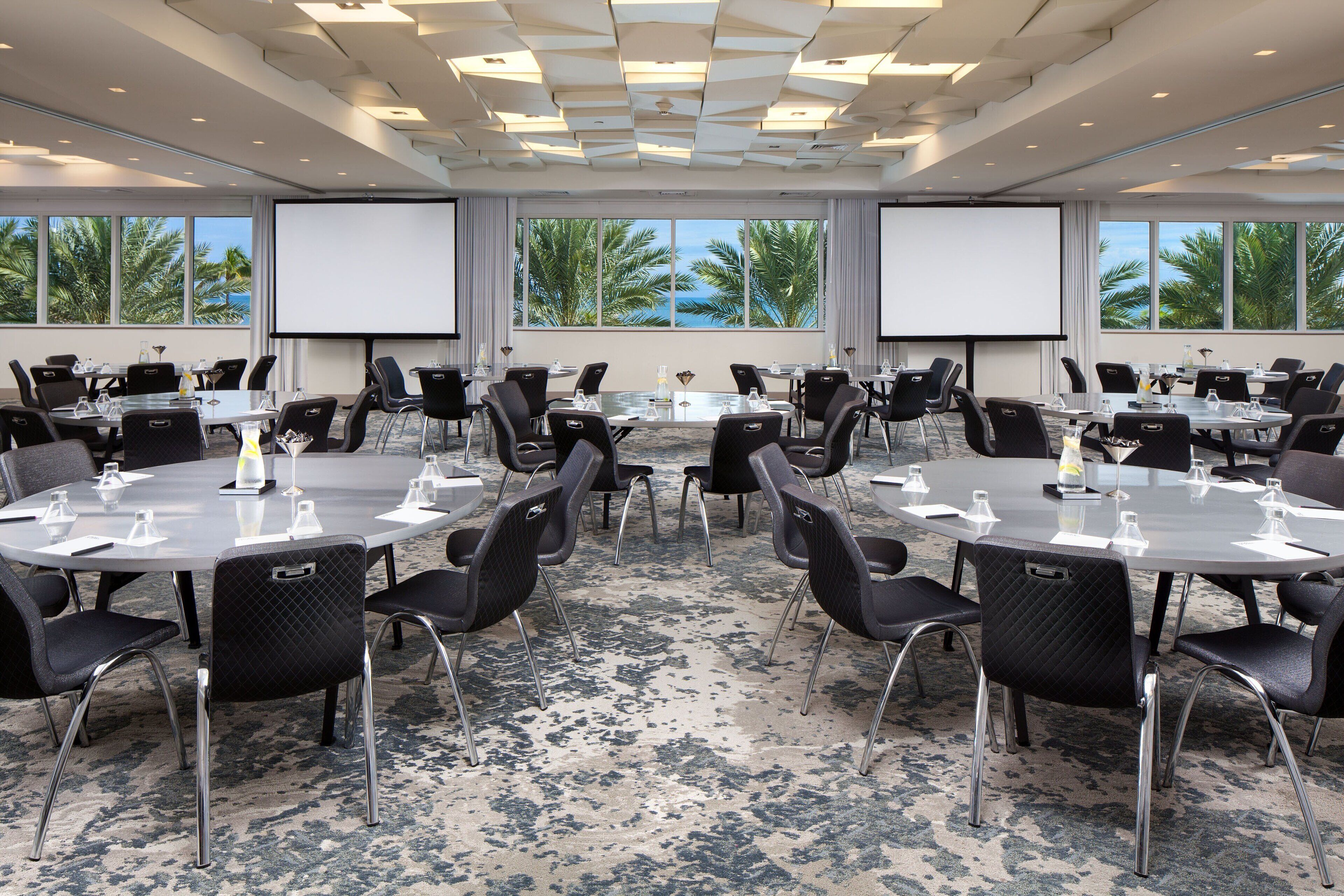 Un salón de conferencias con varias mesas y sillas, ventanas amplias que dan a vistas de palmeras y al océano, y una pantalla de proyector.