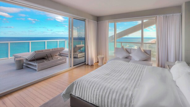 A spacious balcony with a view of the ocean, a large bed with white bedding, and a seating area with cushions. - W Fort Lauderdale (Fort Lauderdale)