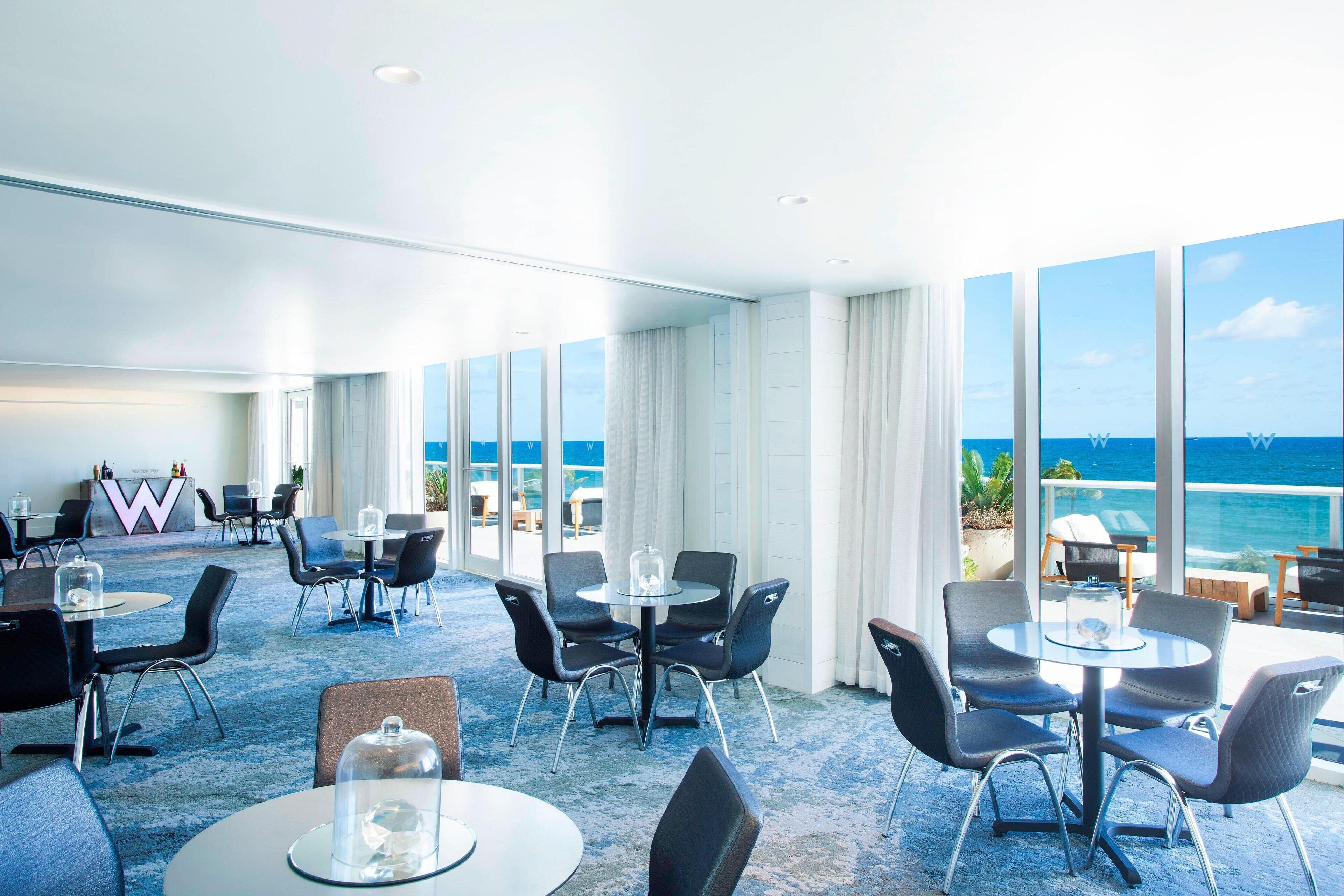 A hotel lobby with round tables and chairs, a reception desk, and large windows offering a view of the ocean.