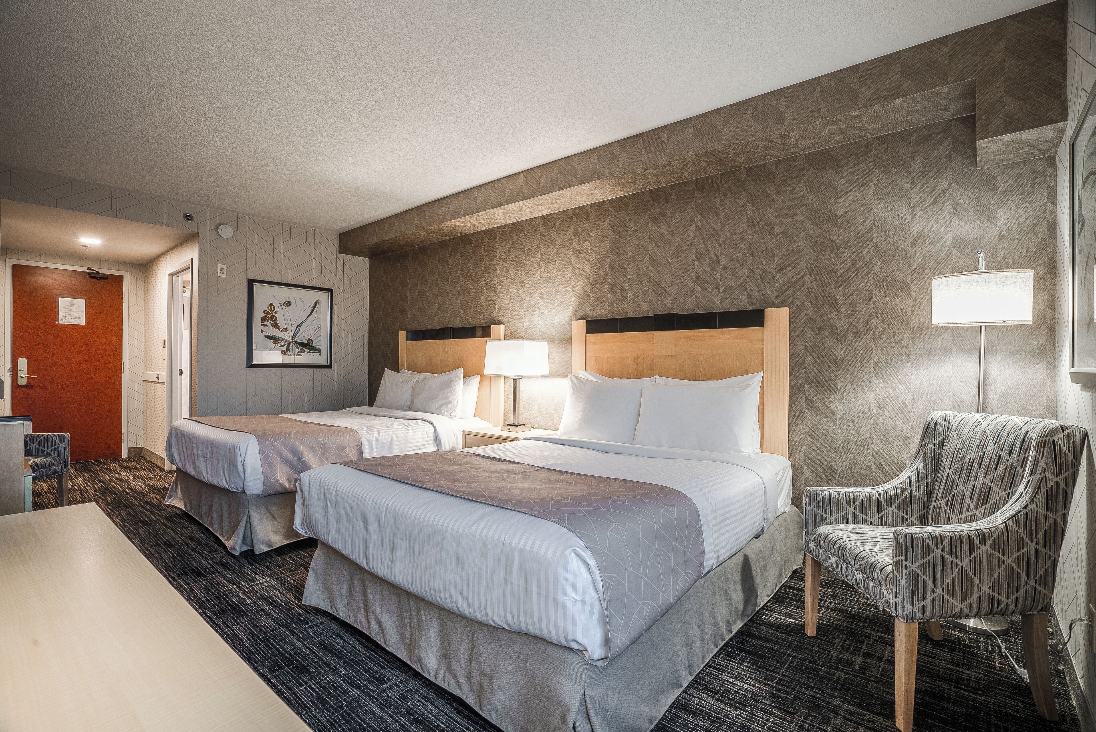 A hotel room with two beds, a desk, a chair, and a lamp.