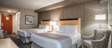 A hotel room with two beds, a desk, a chair, and a lamp.