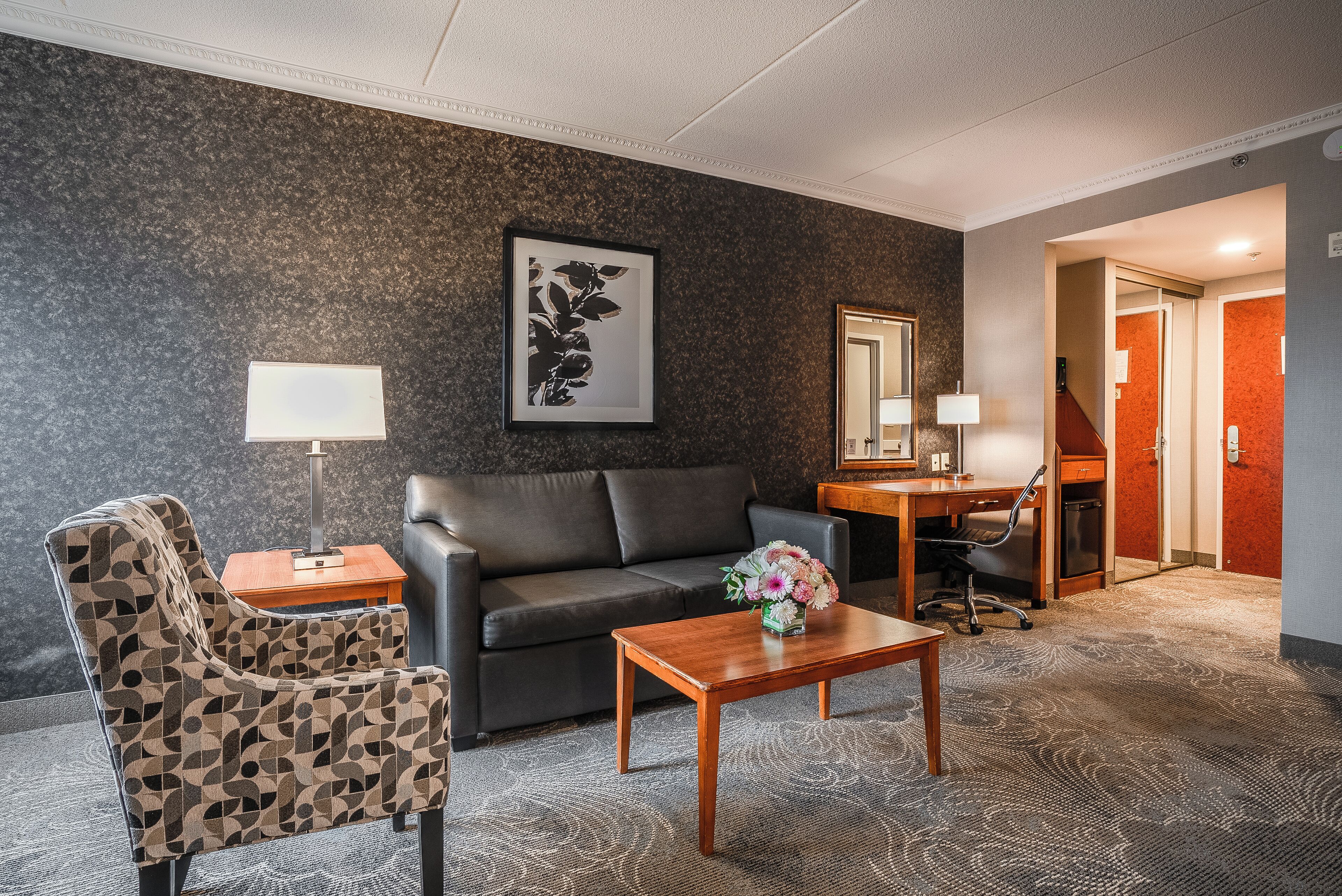 A modern hotel room with a dark patterned wall, a black leather sofa, a patterned armchair, a wooden coffee table with a vase of flowers, a desk with a lamp, and a carpeted floor.