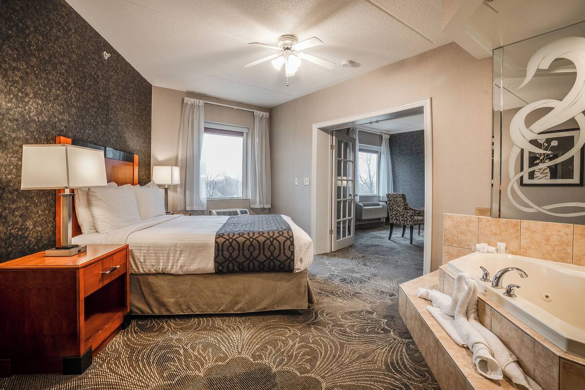 A hotel room with a bed, bedside table, lamp, and a large bathroom with a bathtub and mirror.