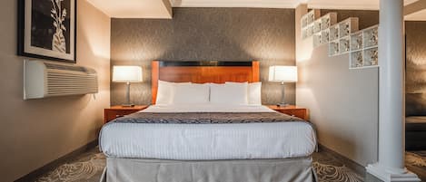 A hotel room with a large bed, two bedside tables, a wall-mounted air conditioner, a framed picture, and a patterned carpet.