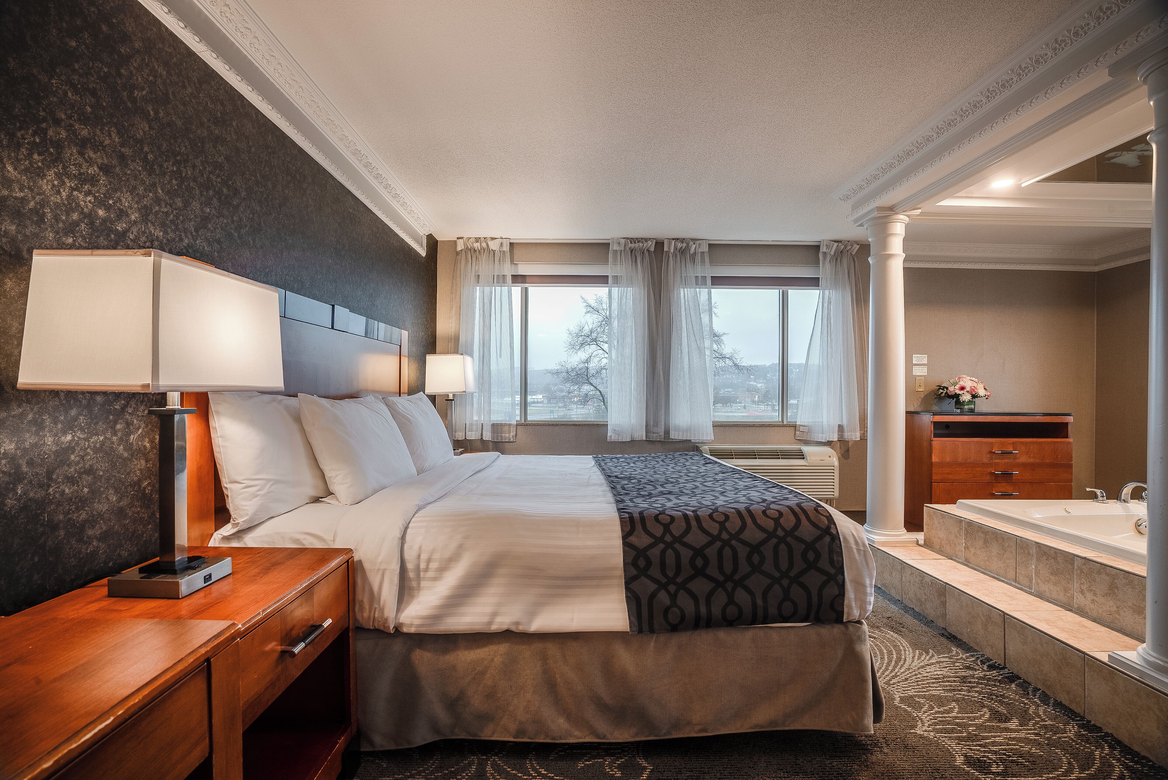 Une chambre d’hôtel avec un grand lit, des lampes de chevet, une table de nuit en bois, une salle de bain avec baignoire et une vue sur la ville par la fenêtre.