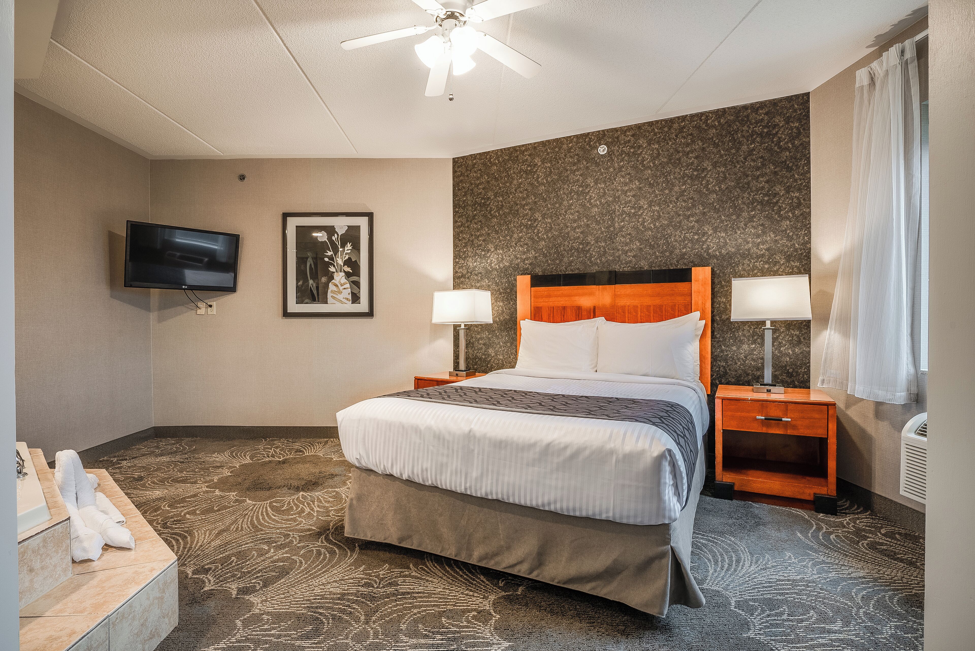 A hotel room with a bed, two bedside tables with lamps, a flat-screen TV mounted on the wall, a framed artwork above the bed, and a patterned carpet.