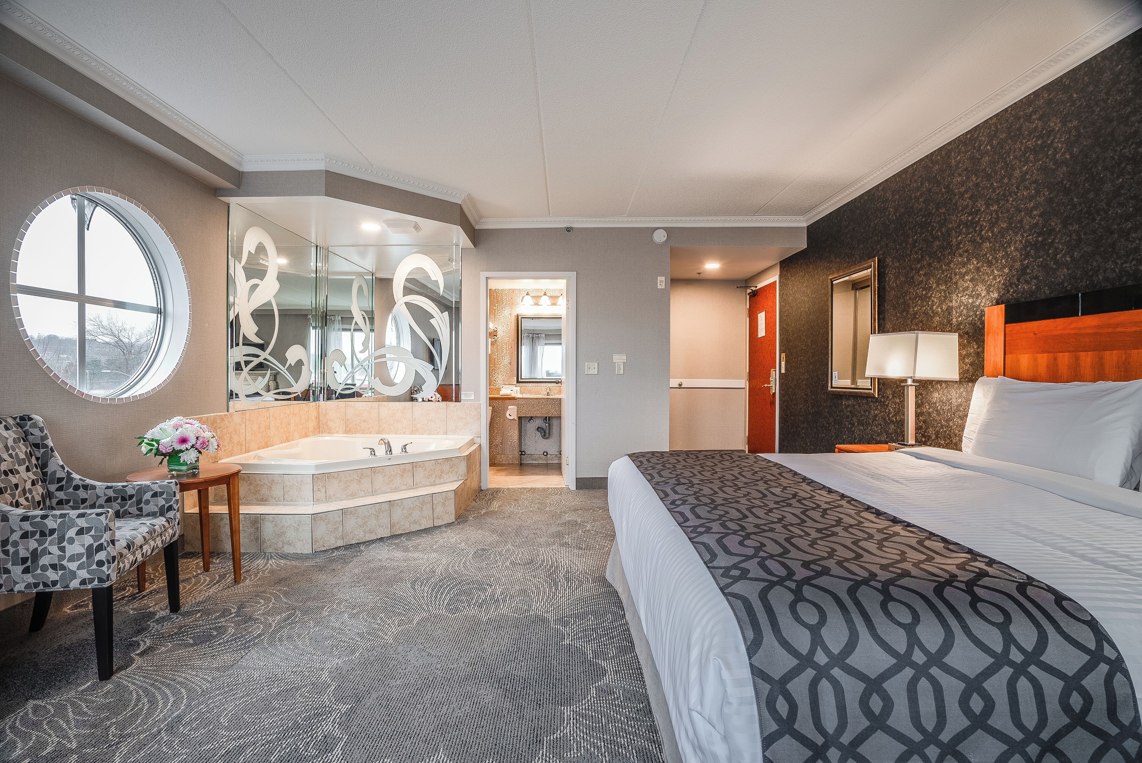 A hotel room with a large bed, a sitting area with a chair, a round window, a bathtub, and a bathroom with a mirror and sink.