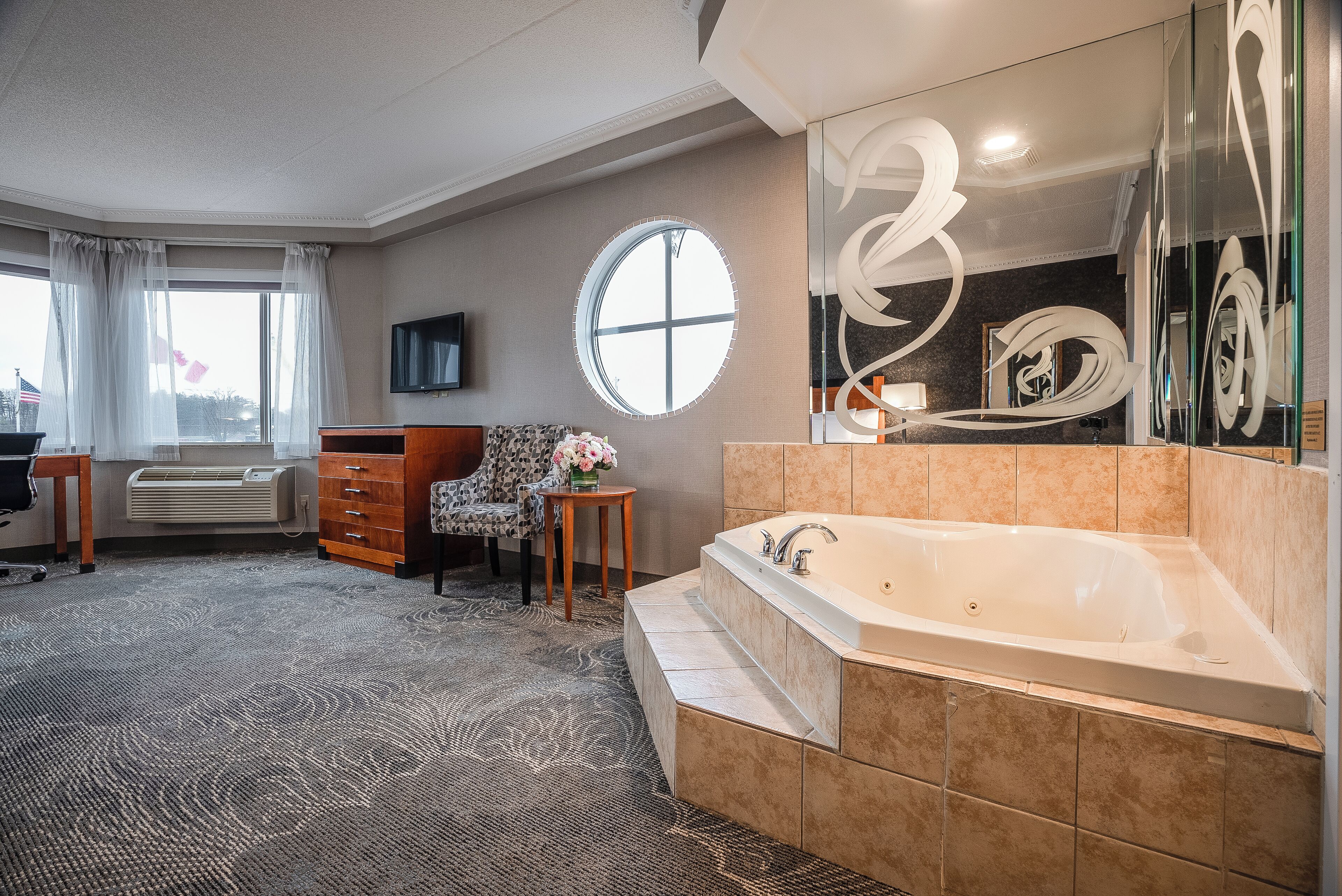 A hotel room with a large bathtub, a round mirror, a desk with a chair, and a view of the outdoors.