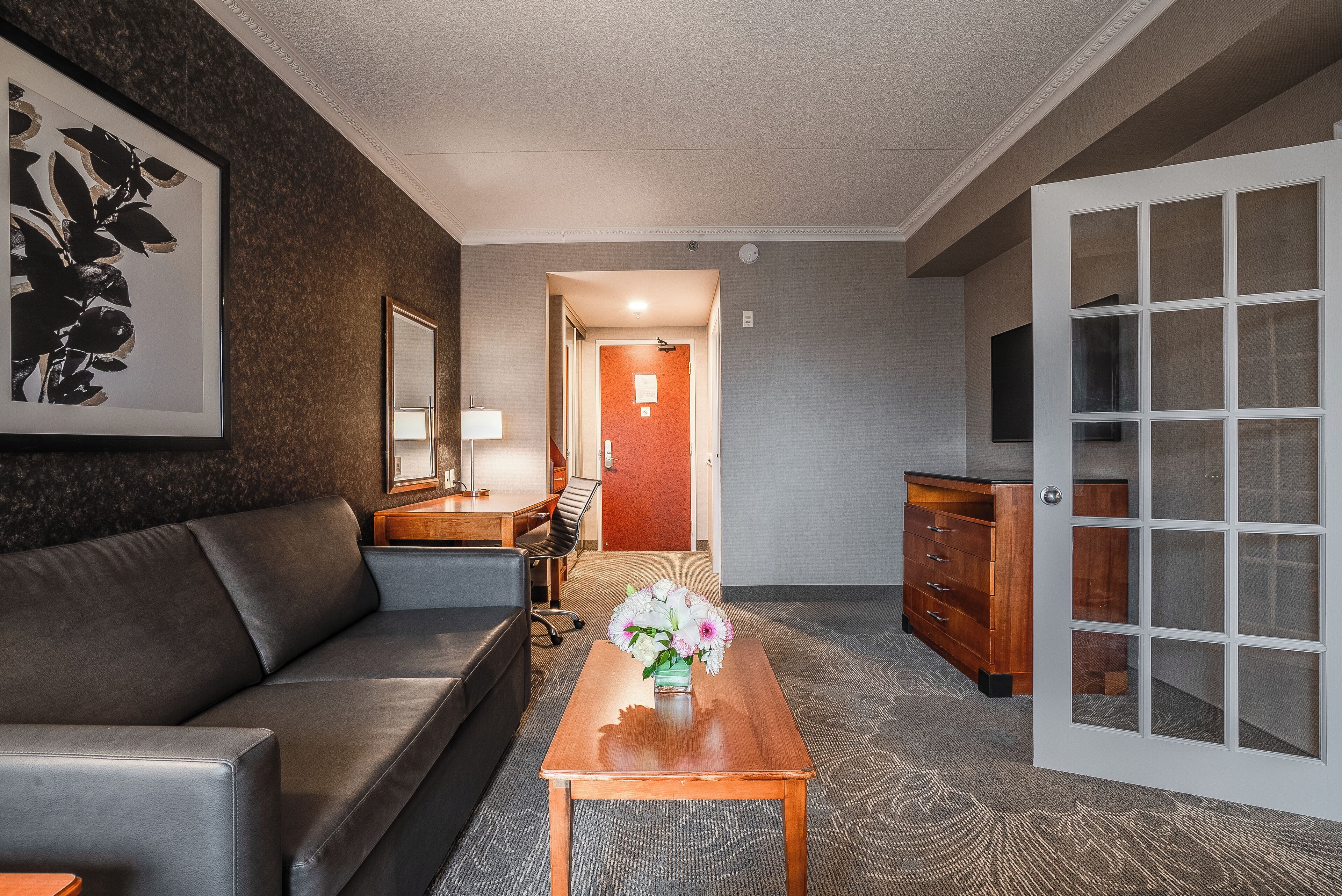 Et hotelværelse med en grå sofa, et træbord med en blomstervase, et skrivebord med stol, en kommode og et fjernsyn på væggen.