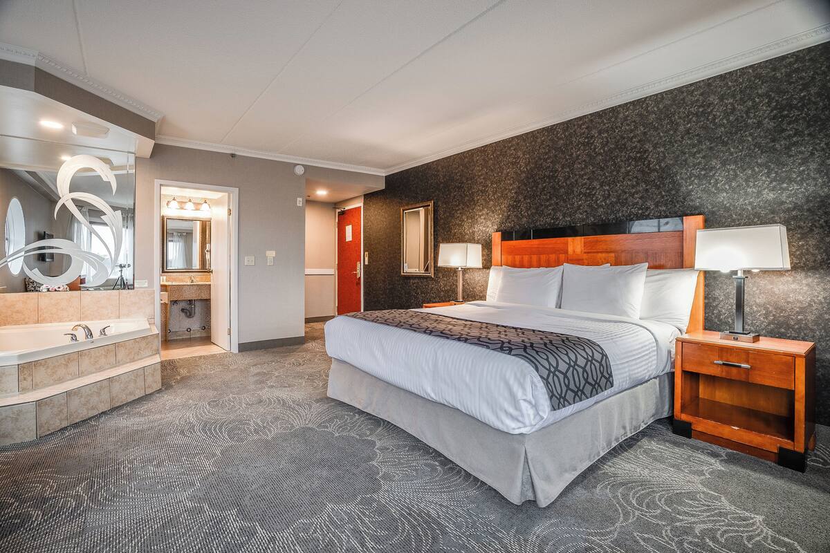A hotel room with a large bed, a wooden nightstand, a mirror, a bathroom with a bathtub, and a patterned carpet.