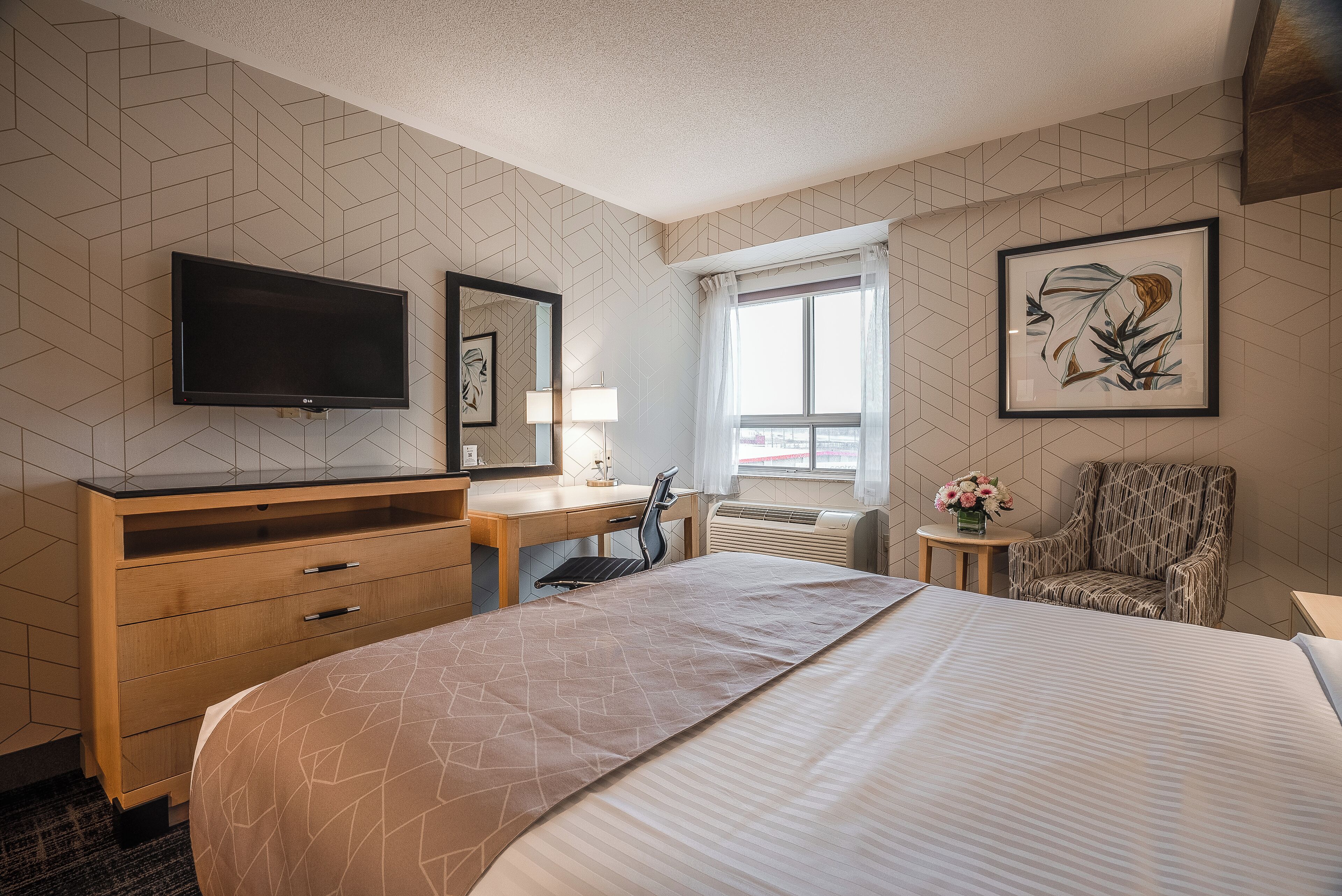 A hotel room with a bed, a desk, a chair, a television, a window with curtains, and a framed picture on the wall.