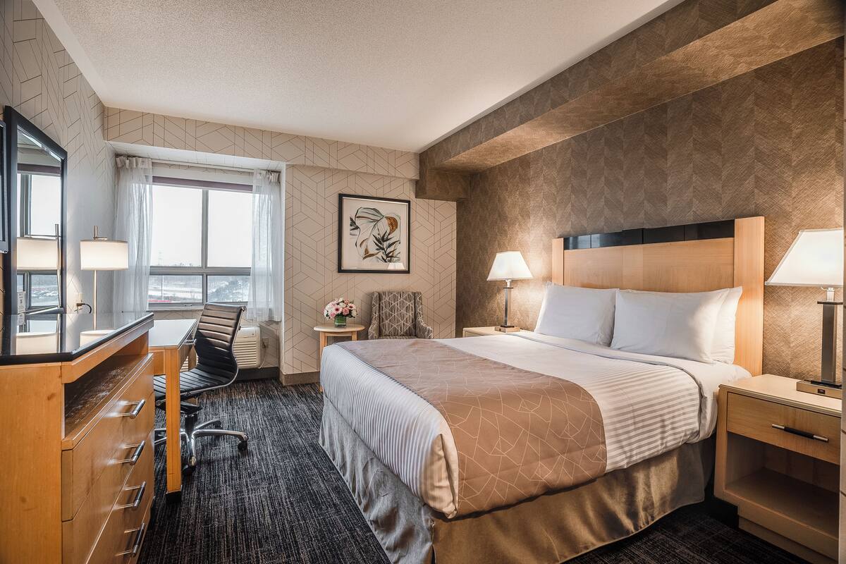 A hotel room with a large bed, a desk with a chair, a dresser, a window with curtains, and a framed artwork on the wall.