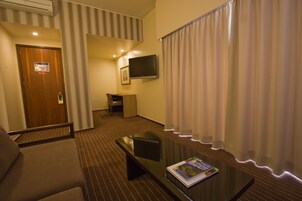 A hotel room with a sofa, a coffee table with a magazine, a TV mounted on the wall, and a door with a sign.
