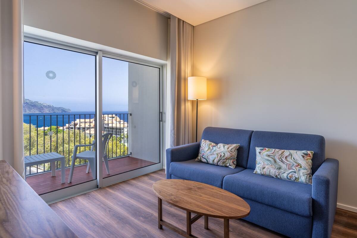 Balkon med havudsigt, en blå sofa med mønstrede puder, et lille træbord og en gulvlampe.