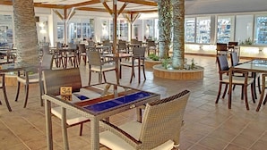 A spacious indoor dining area with wicker furniture and a central table, surrounded by large windows and palm trees. - Sahara Beach Aquapark (Monastir)