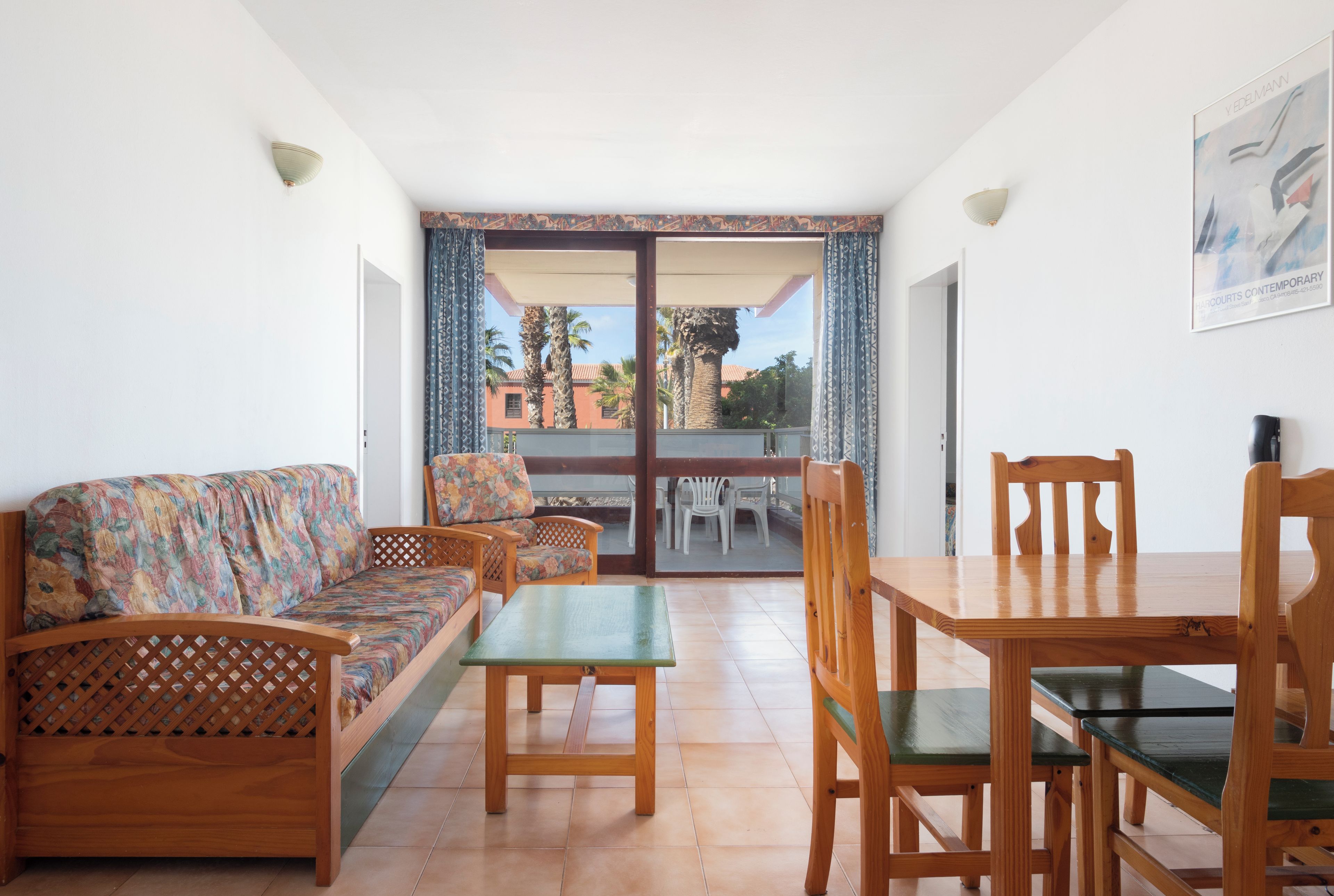 A room with a sofa, a dining table, and chairs, a balcony with a view of palm trees and buildings, and a painting on the wall.