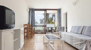 Una habitación con televisión, un sofá, una mesa de comedor con sillas y un balcón con cortinas.