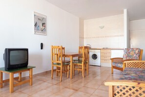 Un espacio habitable funcional con una mesa de comedor, dos sillas de madera, un televisor sobre una mesita, una lavadora y un sofá con un estampado.