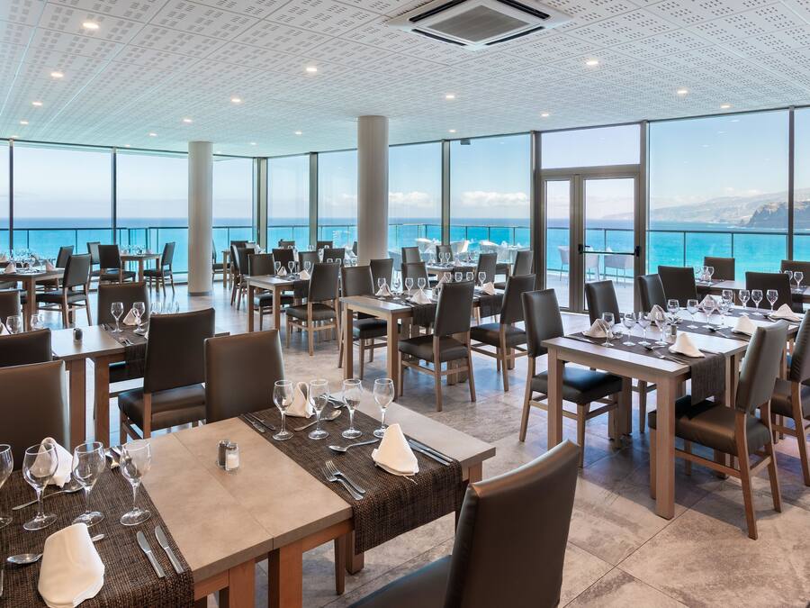 A restaurant with tables set for dining, large windows offering a view of the ocean, and a modern ceiling design.